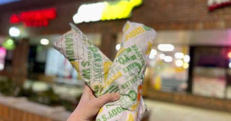 A hand holding two subway footlong sandwiches inside of the subway store