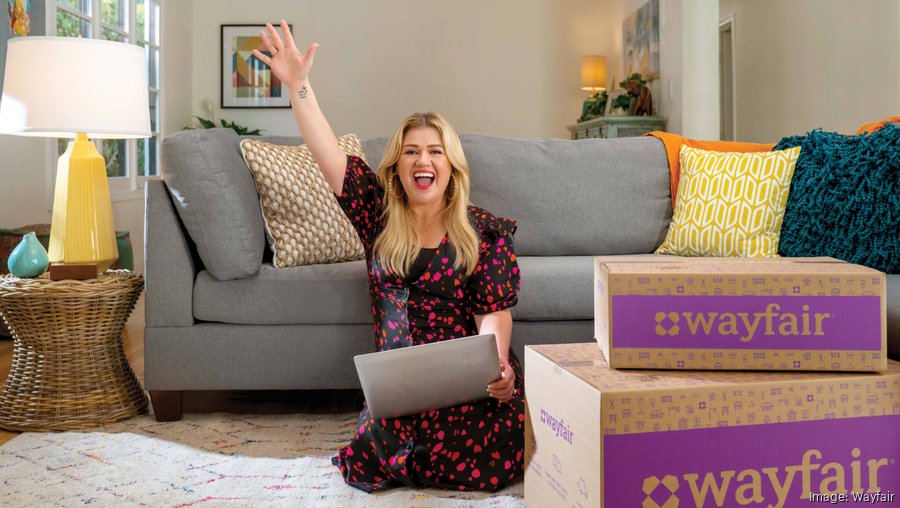 Kelly Clarkson sitting on the floor next to Wayfair boxes and a couch while holding a laptop