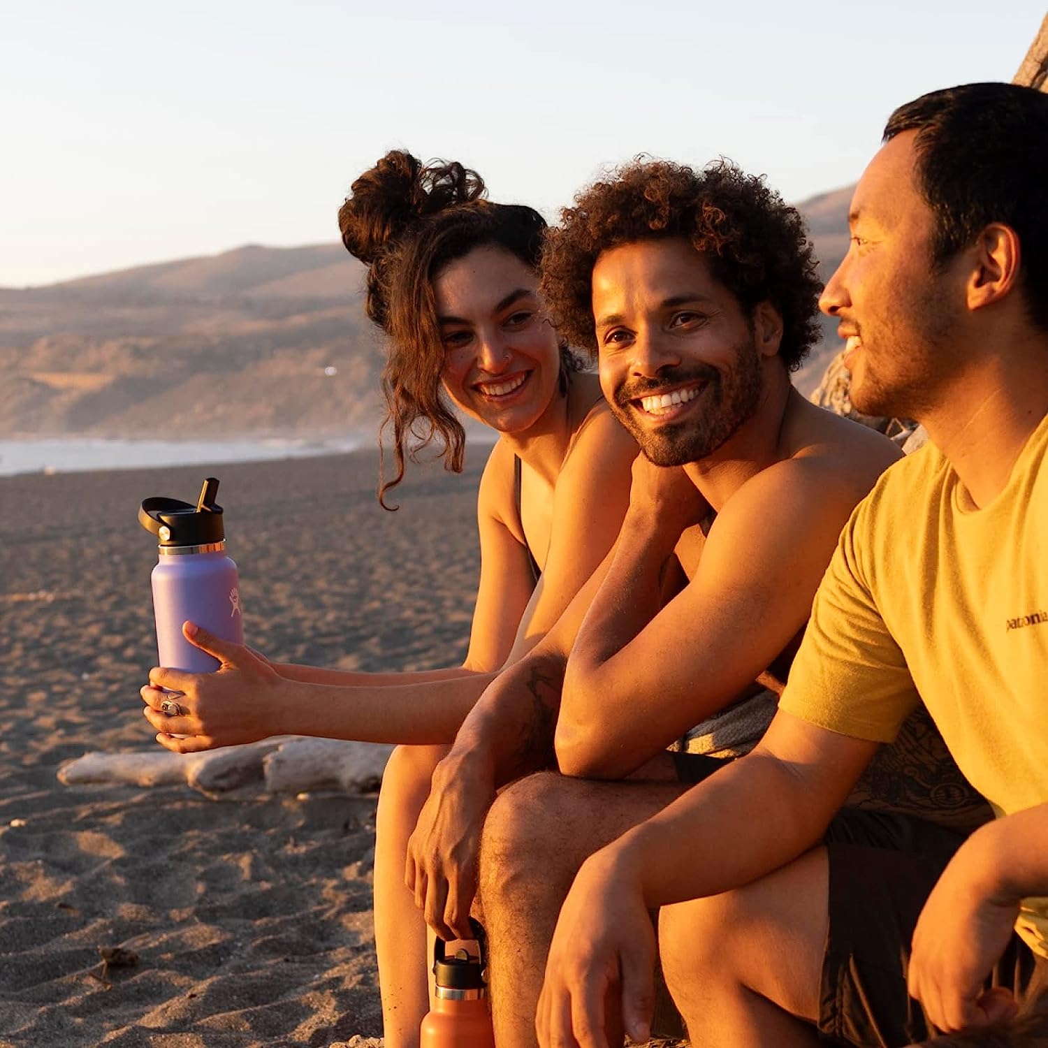 three people sitting on the beach, one of them is holding a water bottle