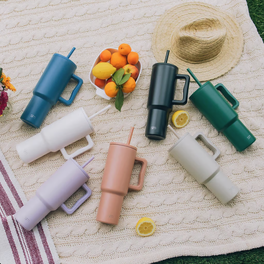 Simple Modern tumblers laid out on a blanket around a bowl of fruit and a straw hat